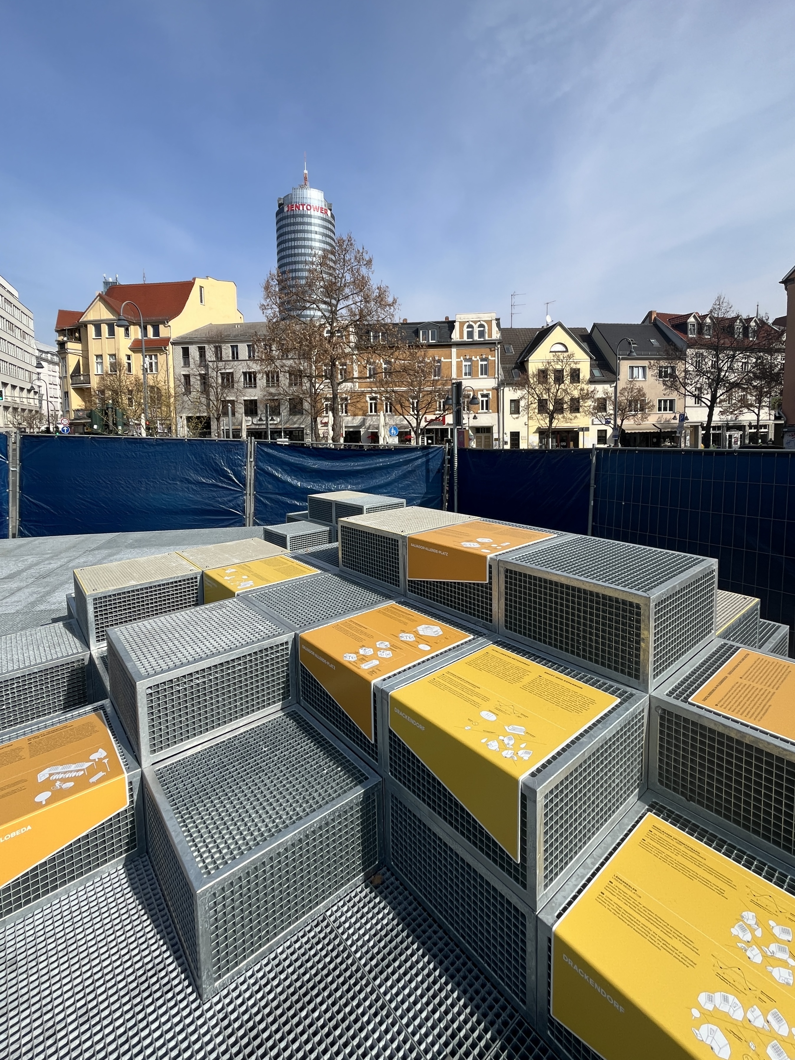 Im Fordergrund sieht man aufeinandergestapelte Metallkonstruktion auf denen Informationstafeln angebracht sind.